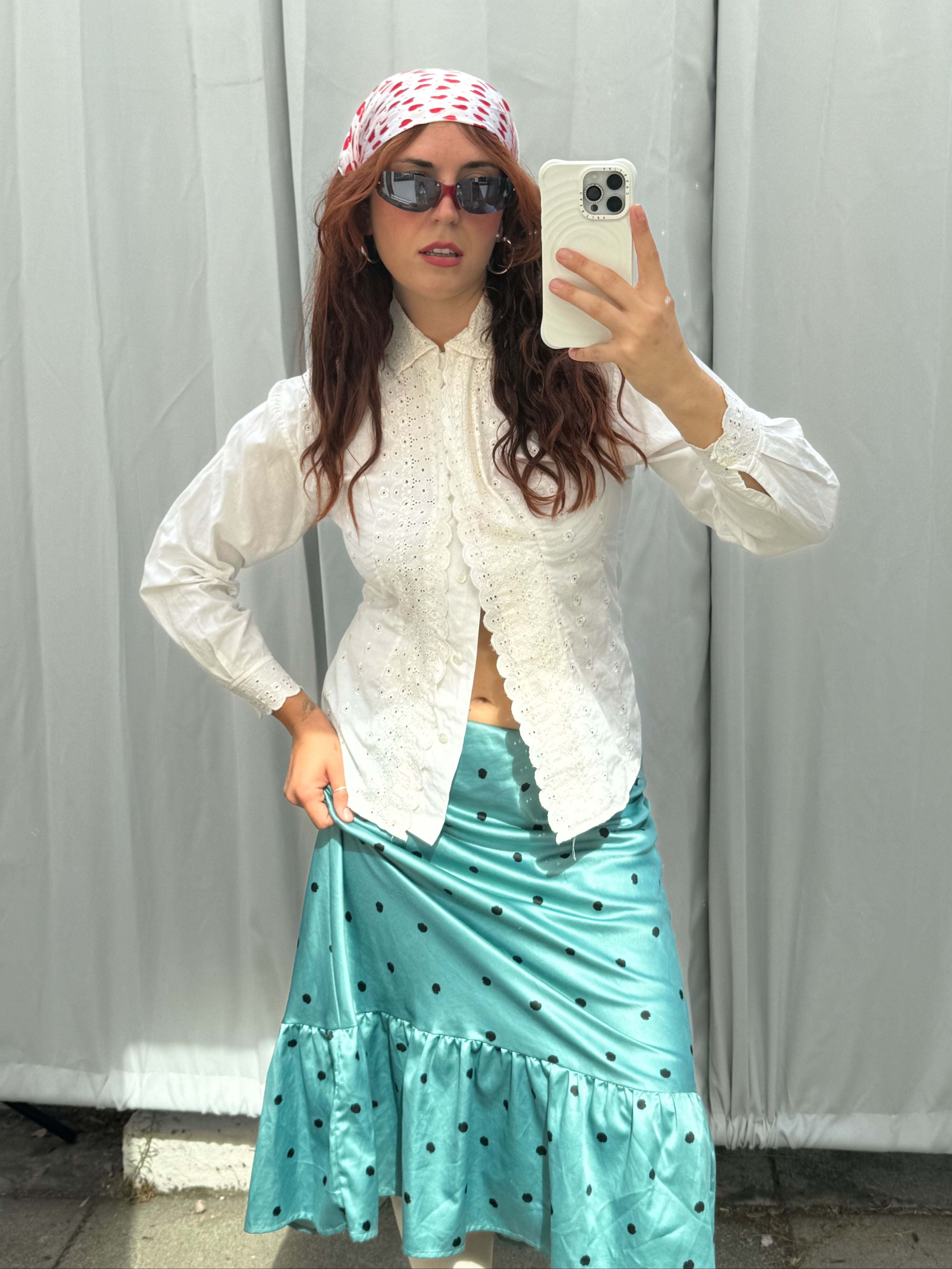 Person taking a mirror selfie wearing a white blouse, teal skirt with polka dots, and sunglasses against a plain background.