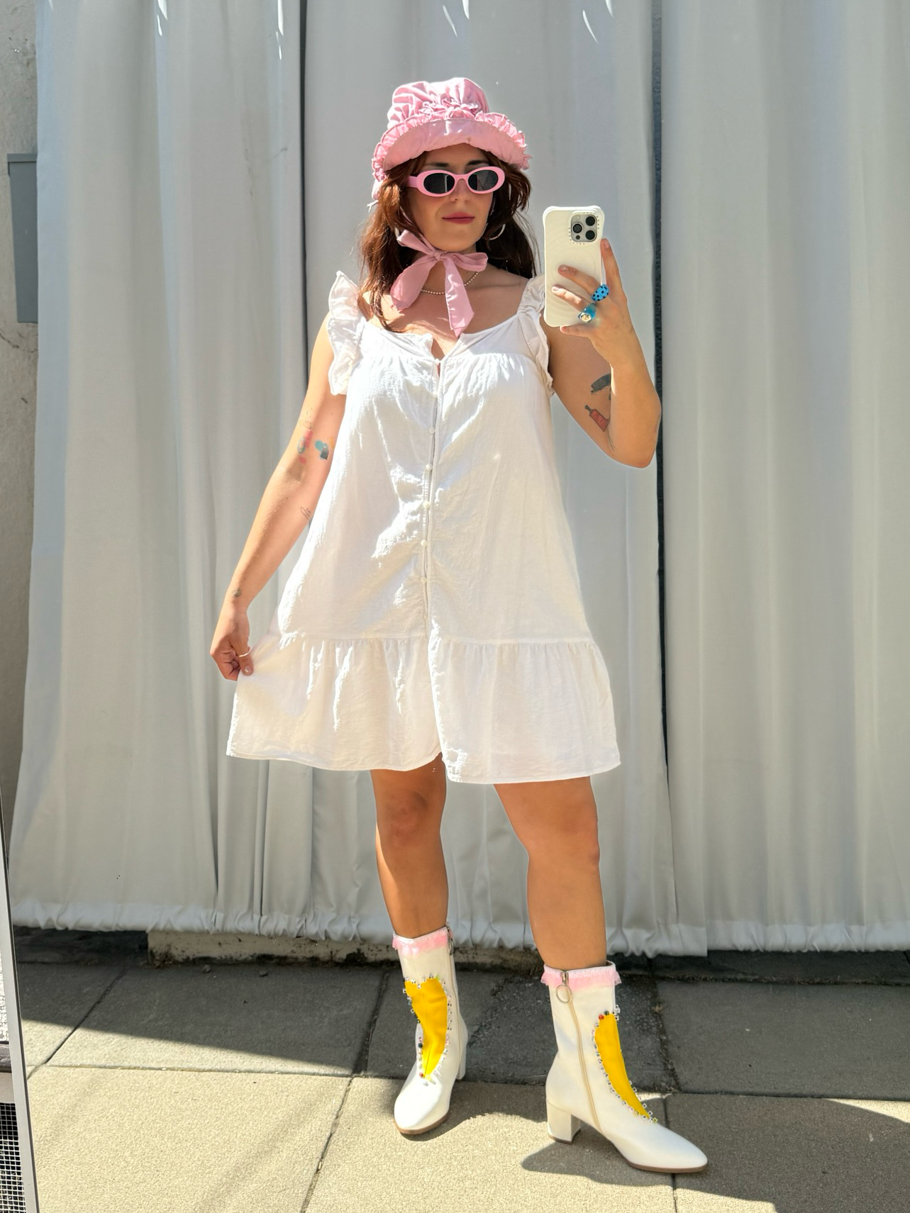 Person taking a mirror selfie wearing a white dress, yellow rain boots, and a pink hat.